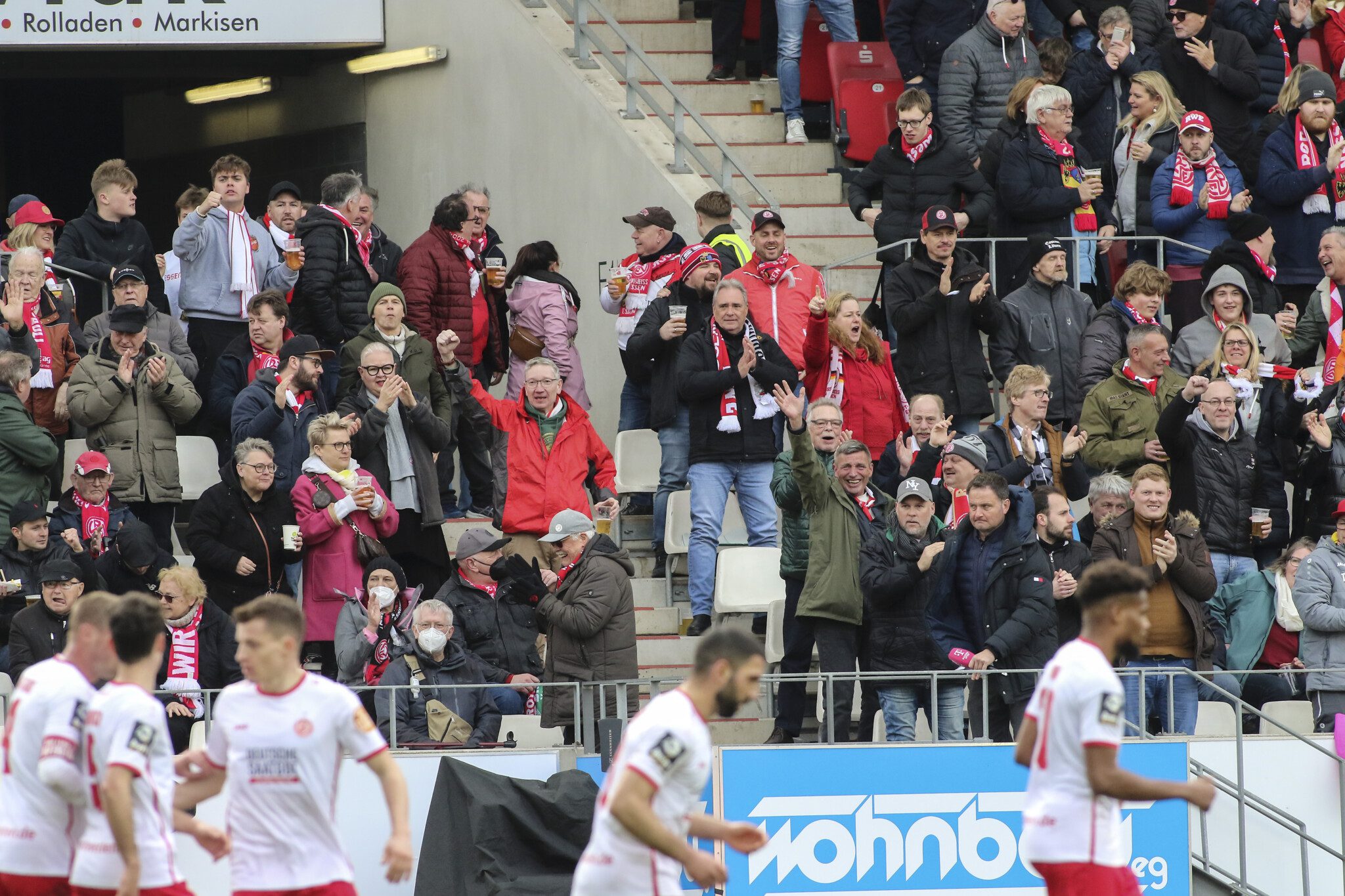 Die Dauerkartenwette 2023/24 ist am Start: Zahlreiche Fans jubeln der RWE-Mannschaft (verschwommen auf dem Foto im Vordergrund) zu. Im Hintergrund sind Treppen und ein Block-Mundloch zu erkennen.